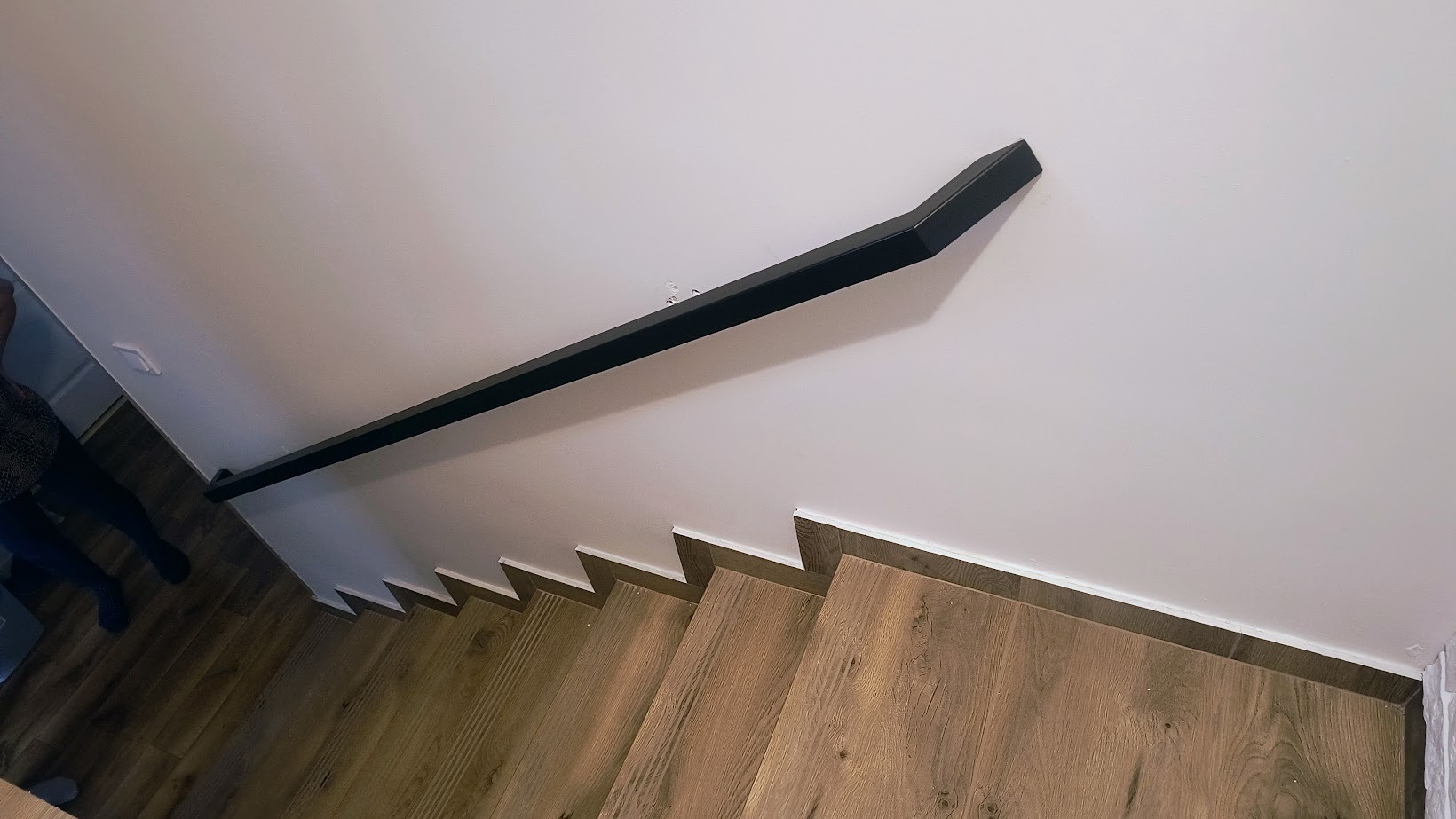 Czarna, metalowa poręcz zamocowana do ściany przy schodach z drewnianymi stopniami, widok z góry.