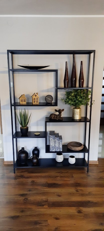 Czarny, metalowy regał z asymetrycznie rozmieszczonymi półkami, na których stoją dekoracje: wazony, figurki, rośliny i książki. Regał stoi na brązowej, drewnianej podłodze.