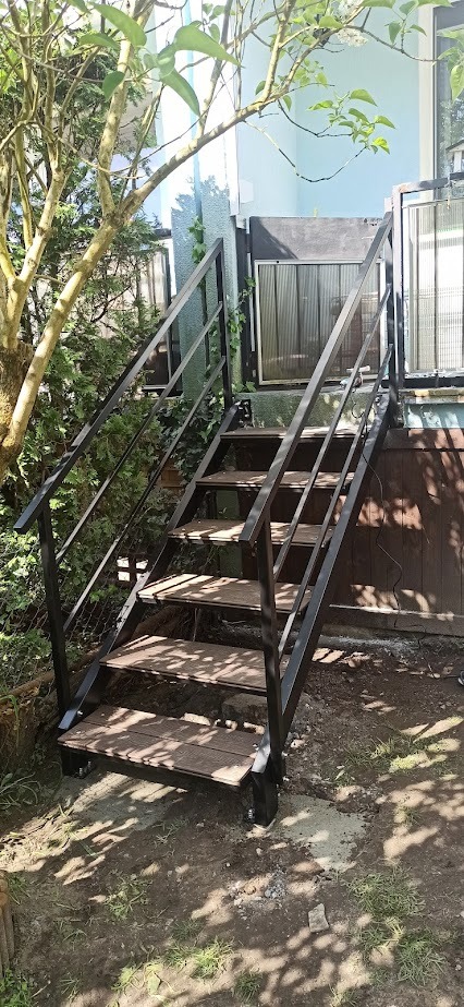 Zewnętrzne schody z czarną metalową ramą i drewnopodobnymi stopniami, prowadzące do wejścia domu, otoczone zielenią.