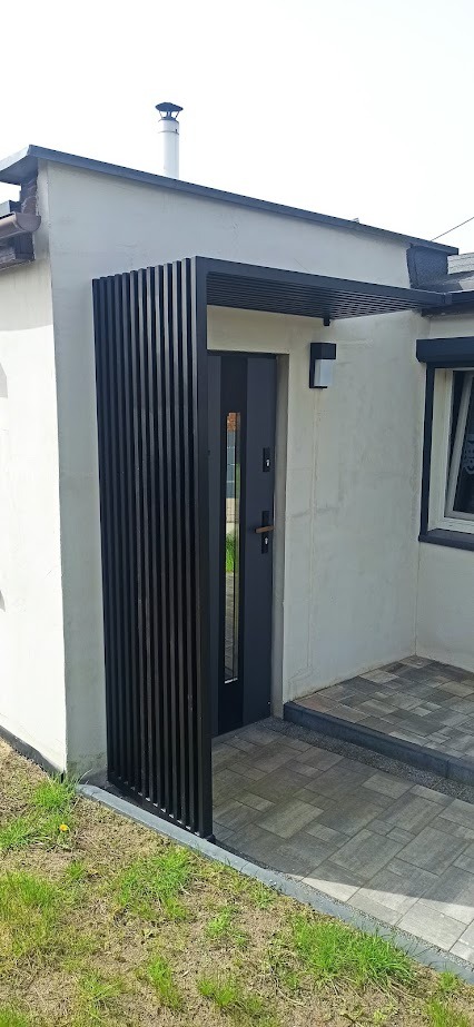 Nowoczesne wejście do domu z czarną, metalową pergolą nad drzwiami i pionową osłoną z metalowych listew po lewej stronie, uzupełnione oświetleniem zewnętrznym i kostką brukową.