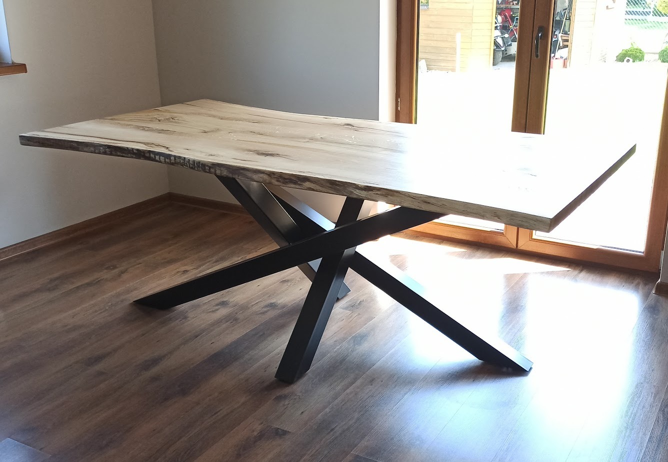 Stół z blatem z jasnego drewna o nieregularnych krawędziach, wsparty na czarnej, metalowej podstawie w kształcie skrzyżowanych belek.