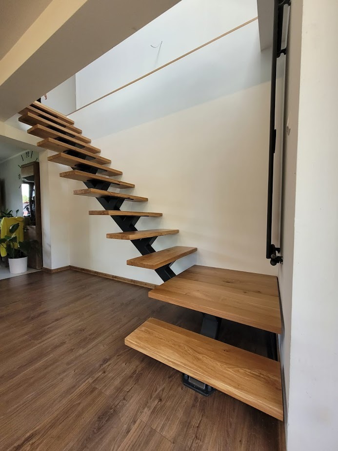 Nowoczesne schody drewniane z metalowymi wspornikami w minimalistycznym wnętrzu. Stopnie z jasnego drewna zamontowane na czarnych, zygzakowatych elementach. Widoczna drewniana podłoga i białe ściany.