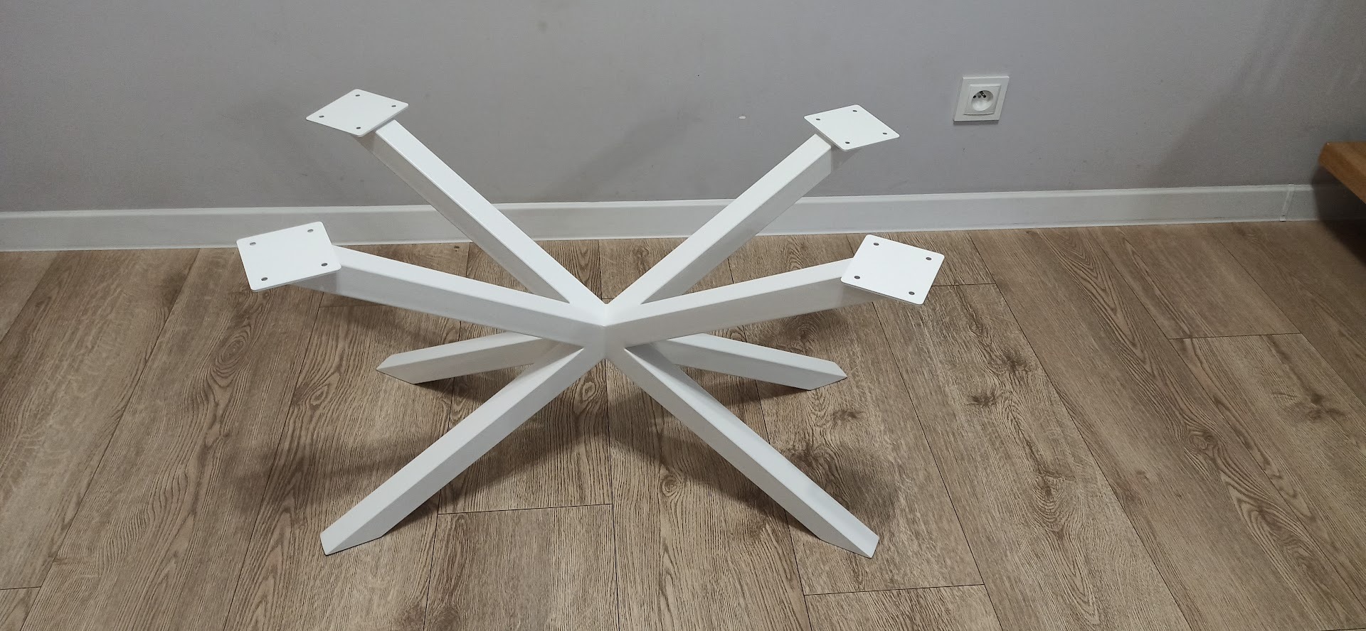 Biała, metalowa podstawa stołu z sześcioma ramionami, ułożona na podłodze z paneli imitujących drewno, widoczne kwadratowe mocowania na blaty.