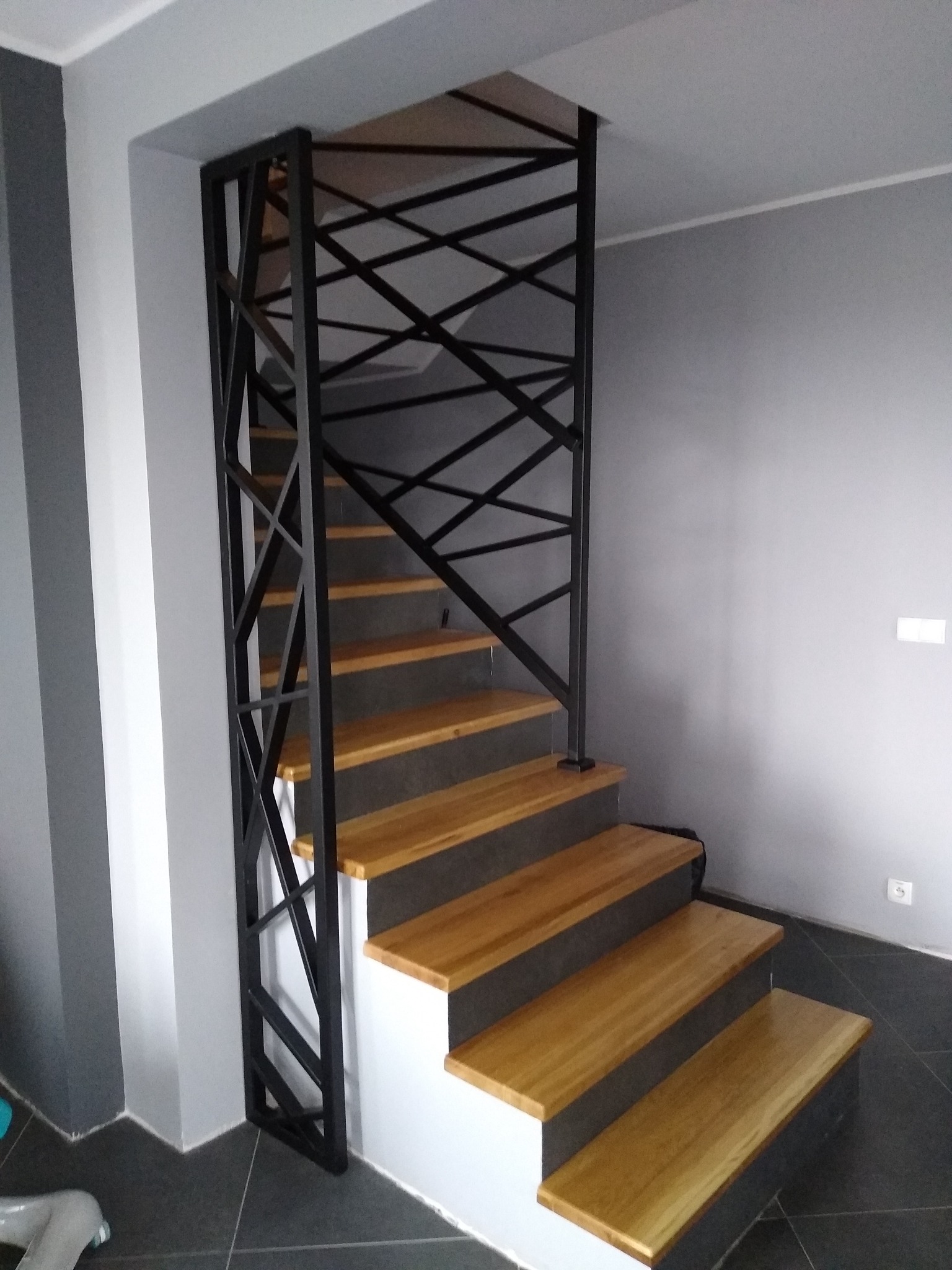 Schody drewniane z metalową, geometryczną balustradą w nowoczesnym wnętrzu z szarą kolorystyką.