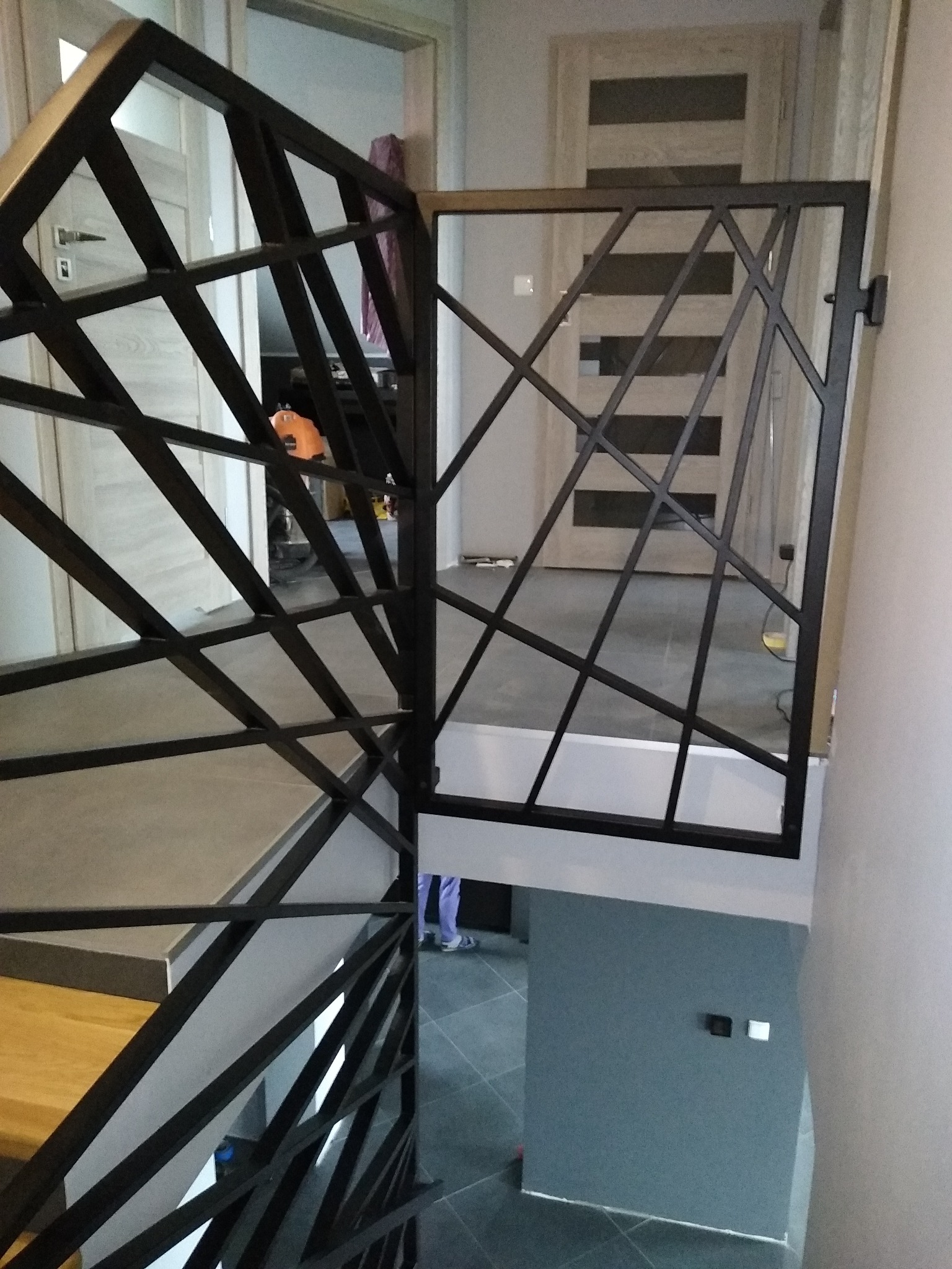 Nowoczesna balustrada schodowa ze stali malowanej proszkowo na czarno, z geometrycznym wzorem rombów i trójkątów, widoczna z góry, z fragmentem drewnianych schodów i korytarzem w tle.