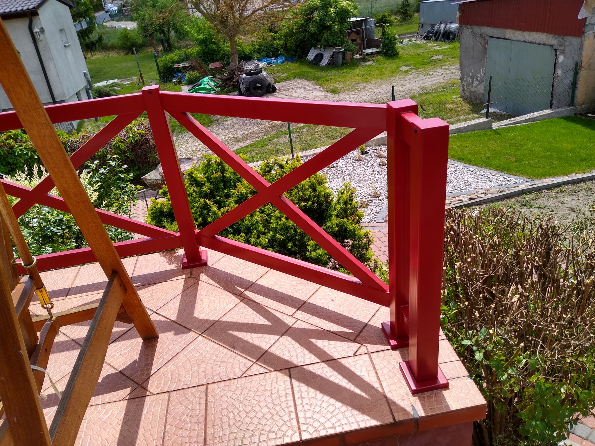 Czerwona balustrada balkonowa z drewnianymi elementami drabiny, widok na ogród z zielenią i garażem w tle.
