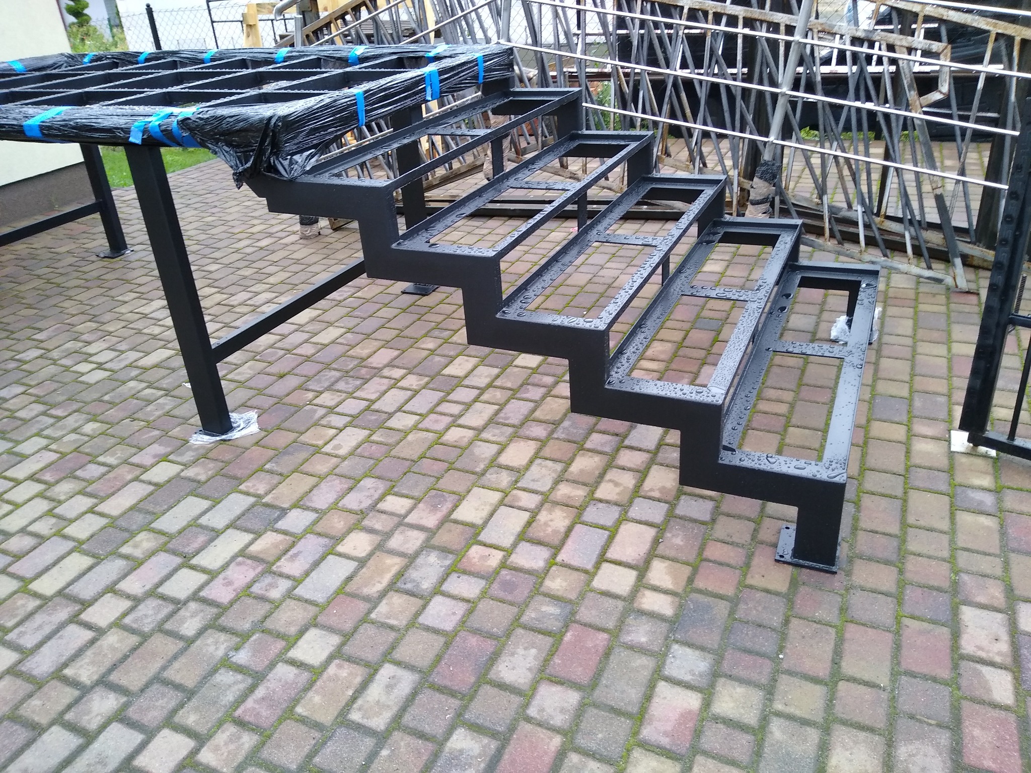 Konstrukcja schodów metalowych, pomalowana na czarno, z kroplami wody, częściowo owinięta folią zabezpieczającą z niebieską taśmą, ułożona na kostce brukowej. W tle elementy balustrady.