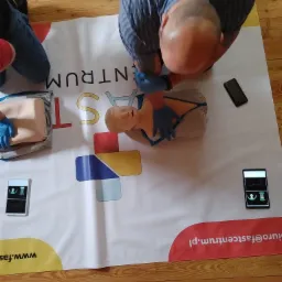 Instruktor w niebieskich rękawiczkach demonstruje uciskanie klatki piersiowej na manekinie podczas kursu pierwszej pomocy, na macie z logo Fastcentrum.pl, obok leży smartfon i tablet.