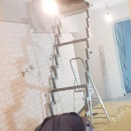 Fragment ściany z cegły pomalowanej na biało, częściowo obłożonej płytami kartonowo-gipsowymi, wspartej na metalowym stelażu, drabina, widoczne kable elektryczne i folia ochronna na podłodze, lampa...