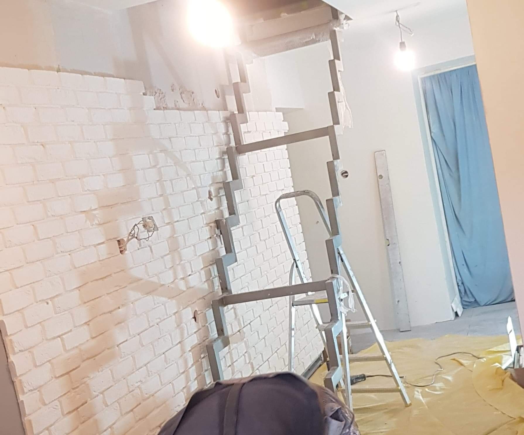 Fragment ściany z cegły pomalowanej na biało, częściowo obłożonej płytami kartonowo-gipsowymi, wspartej na metalowym stelażu, drabina, widoczne kable elektryczne i folia ochronna na podłodze, lampa...