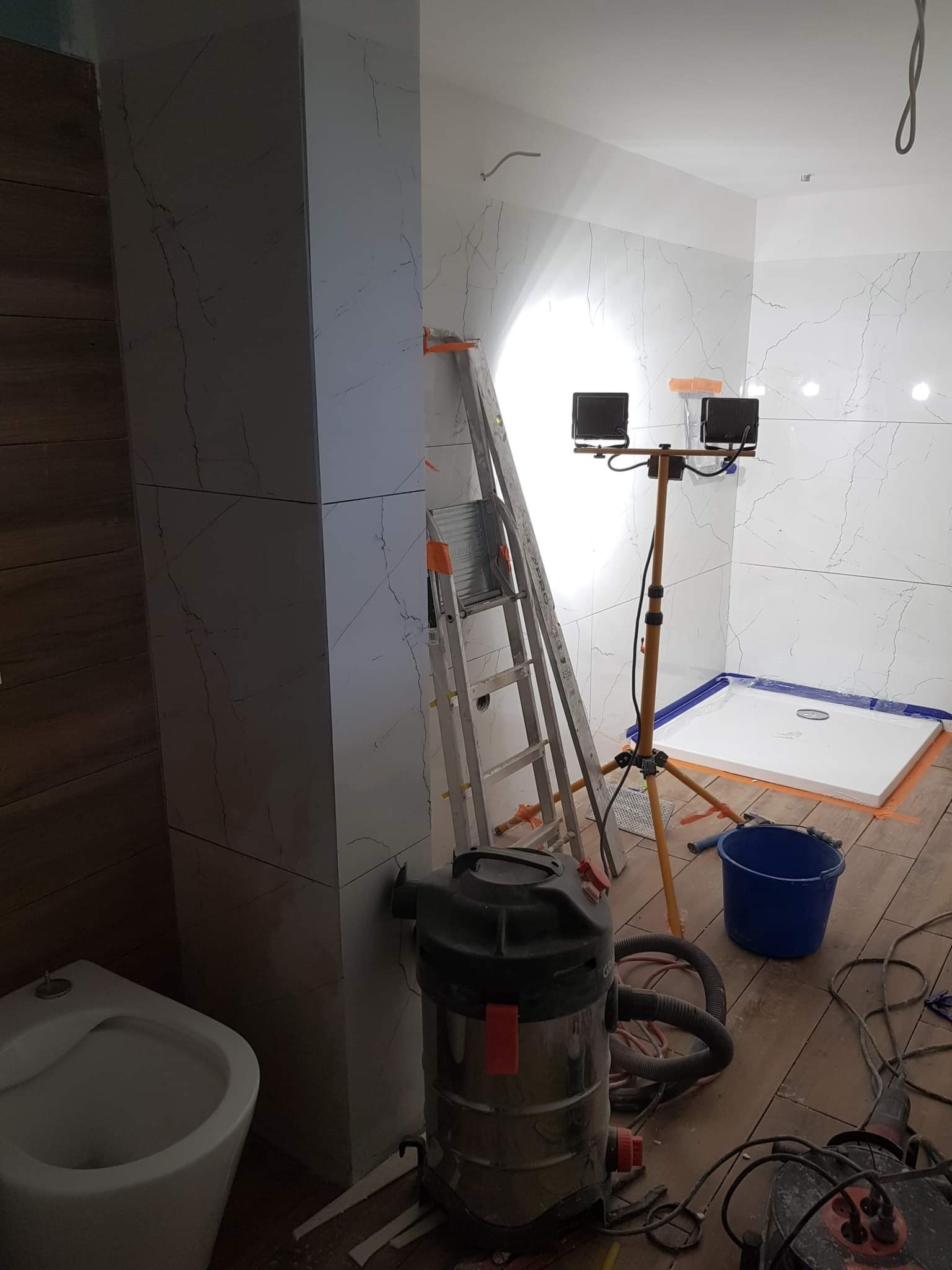 Wnętrze łazienki w trakcie prac wykończeniowych: białe płytki imitujące marmur na ścianach, brodzik zabezpieczony folią, drabina, lampa budowlana na statywie, odkurzacz przemysłowy i niebieskie...