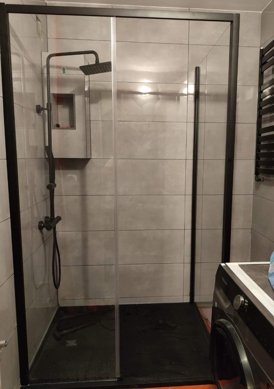 Nowoczesna kabina prysznicowa z czarnymi profilami i deszczownicą w łazience wyłożonej szarymi, poziomymi płytkami, z widoczną pralką w rogu.