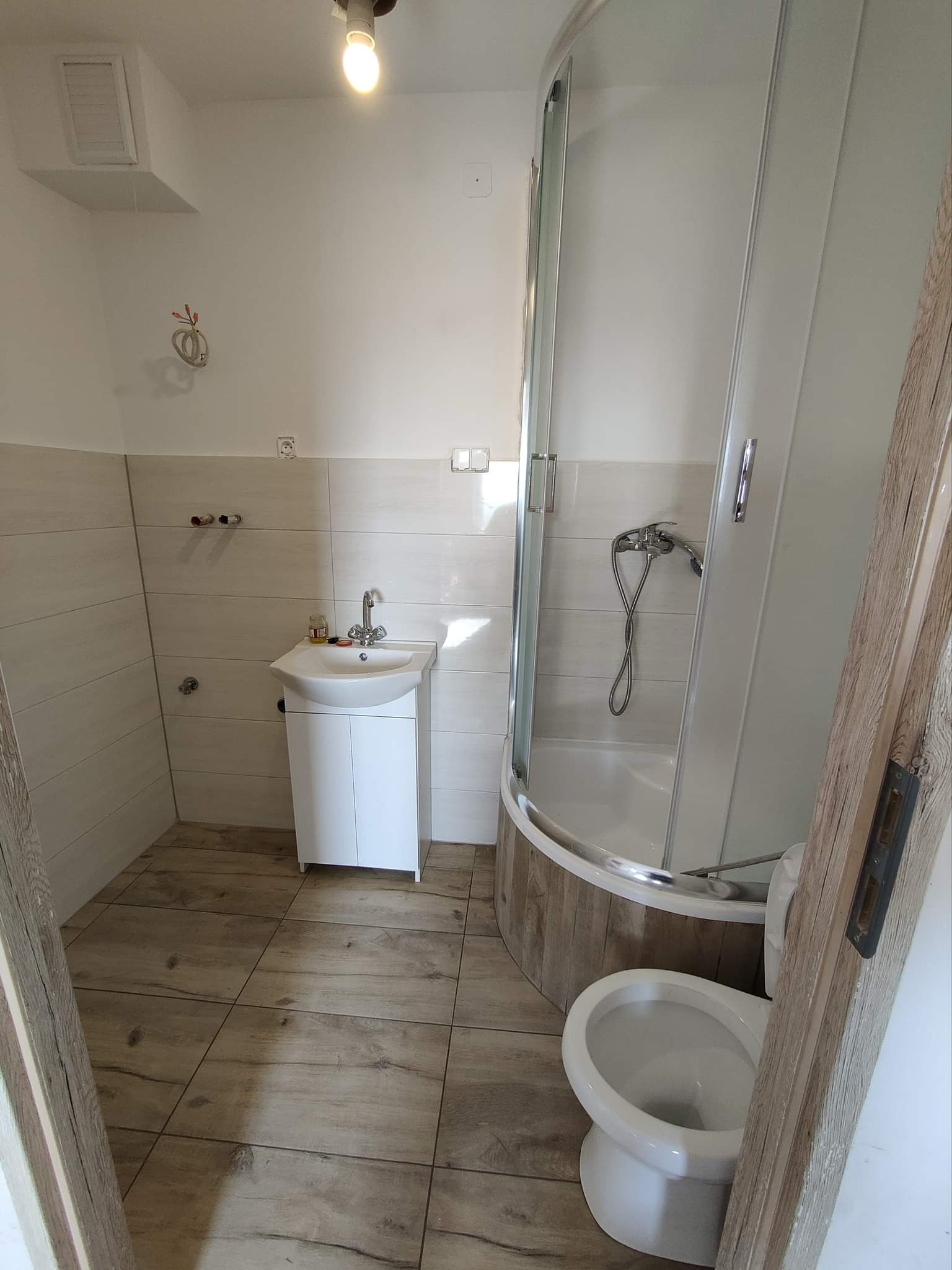 Ukończona łazienka z kabiną prysznicową z zaokrąglonymi szybami, białą szafką pod umywalką, toaletą i płytkami imitującymi drewno na podłodze i częściowo na ścianach kabiny.