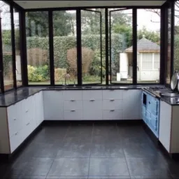 Nowoczesna kuchnia z białymi szafkami, czarnym blatem i niebieskim piecem, otoczona szklanymi ścianami z widokiem na ogr&oacute;d.