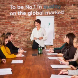 Kobieta w białej koszuli prowadzi prezentację dla grupy osób siedzących przy drewnianym stole; na ścianie z czerwonej cegły widnieje napis 'To be No.1 in quality on the global market!'
