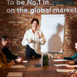 Prezentacja na tle ceglanej ściany z napisem 'To be No.1 in quality on the global market!'. Kobieta w białej koszuli omawia zagadnienie 'JAK?' zapisane na flipcharcie, uczestnicy szkolenia robią...