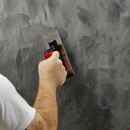 VIP Interior Decor - Nakładanie dekoracyjnej szarej masy na ścianę za pomocą pacy z czerwoną rączką, widoczny fragment białej koszulki osoby wykonującej pracę.