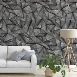 Czarno-biała, geometryczna tapeta z motywem trójkątów, uzupełniona białym narożnikiem, szarymi poduszkami, lampą stojącą na drewnianym trójnogu i rośliną doniczkową w plecionym koszu.