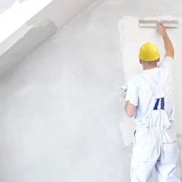 VIP Interior Decor - Malarz w żółtym kasku wygładza ścianę szpachlą w jasnym pokoju na poddaszu, widoczne narzędzia na parapecie.