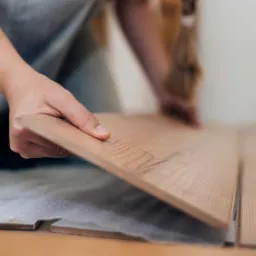 Układanie paneli podłogowych: dłoń dopasowuje panel do już ułożonej podłogi, widoczny karton z panelami i narzędzie w tle.