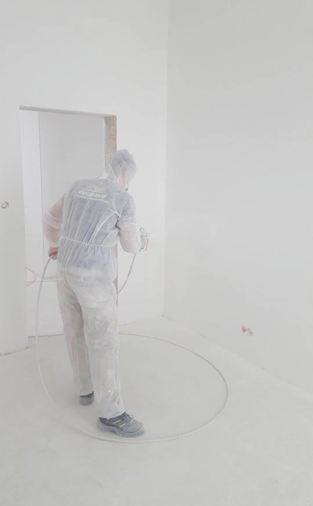 Pracownik w kombinezonie ochronnym maluje ściany agregatem natryskowym w jasnym, pustym pomieszczeniu. Widoczne przewody i niewielkie niedoskonałości na ścianie.