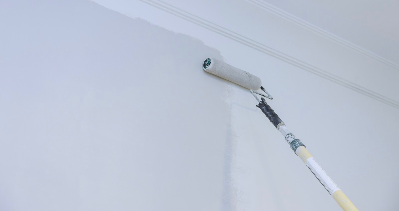 Świeżo pomalowana jasna ściana z widocznym wałkiem malarskim na teleskopowym uchwycie, fragment listwy przysufitowej w tle.