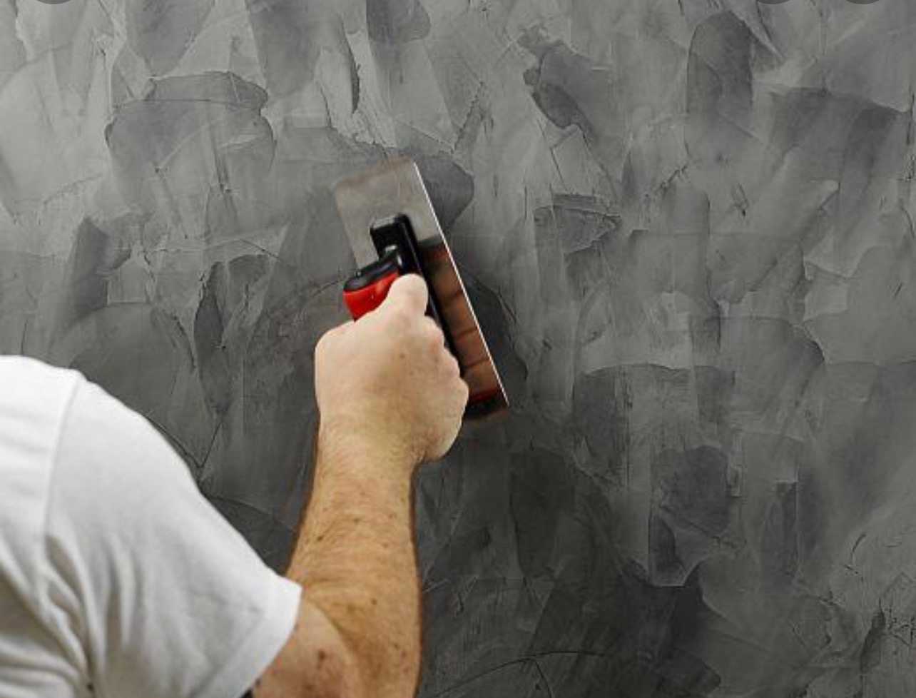 Nakładanie dekoracyjnej szarej masy na ścianę za pomocą pacy z czerwoną rączką, widoczny fragment białej koszulki osoby wykonującej pracę.