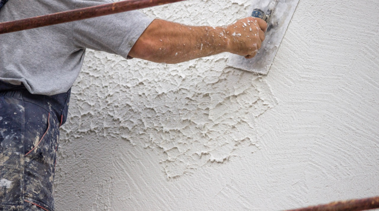 Nakładanie strukturalnego tynku na ścianę przez pracownika w ubrudzonych spodniach roboczych, widoczne rusztowanie.