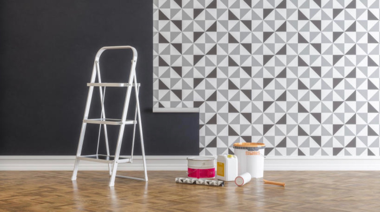 Przygotowanie do tapetowania ściany: drabina aluminiowa, puszki z klejem i farbą, wałek i rolka tapety w geometryczny wzór trójkątów na podłodze z parkietu.
