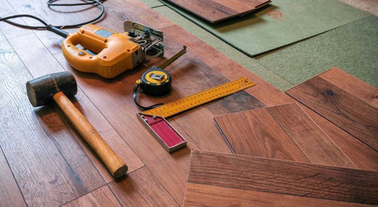 Przygotowanie do układania paneli podłogowych: gumowy młotek, wyrzynarka, miarka i kątownik stolarski leżą na ułożonych panelach z widocznym podkładem.