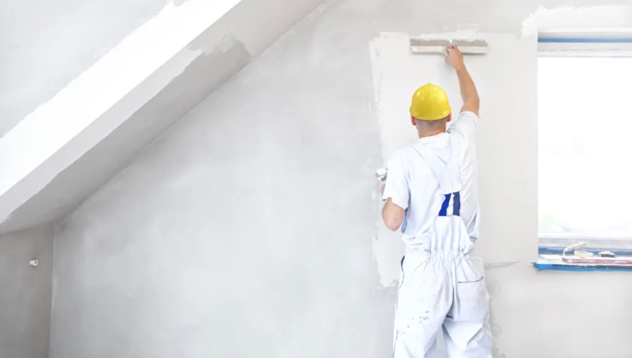 Malarz w żółtym kasku wygładza ścianę szpachlą w jasnym pokoju na poddaszu, widoczne narzędzia na parapecie.