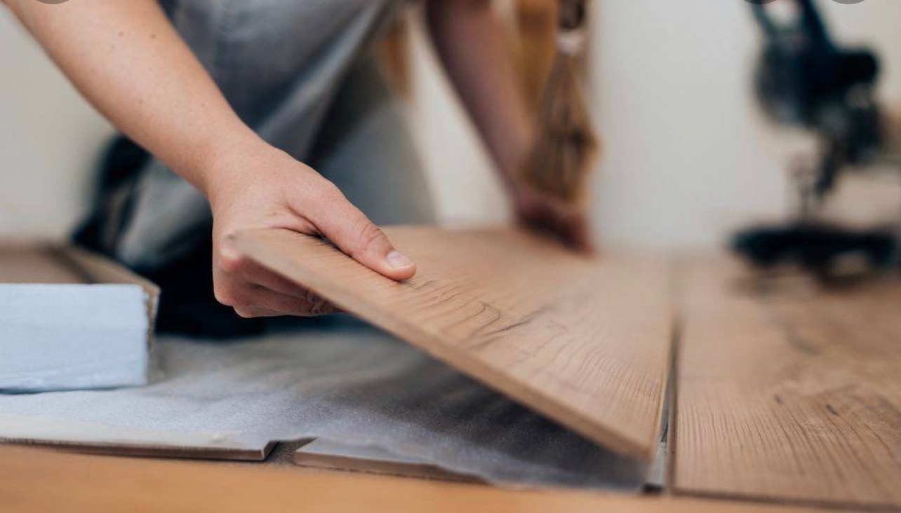 Układanie paneli podłogowych: dłoń dopasowuje panel do już ułożonej podłogi, widoczny karton z panelami i narzędzie w tle.