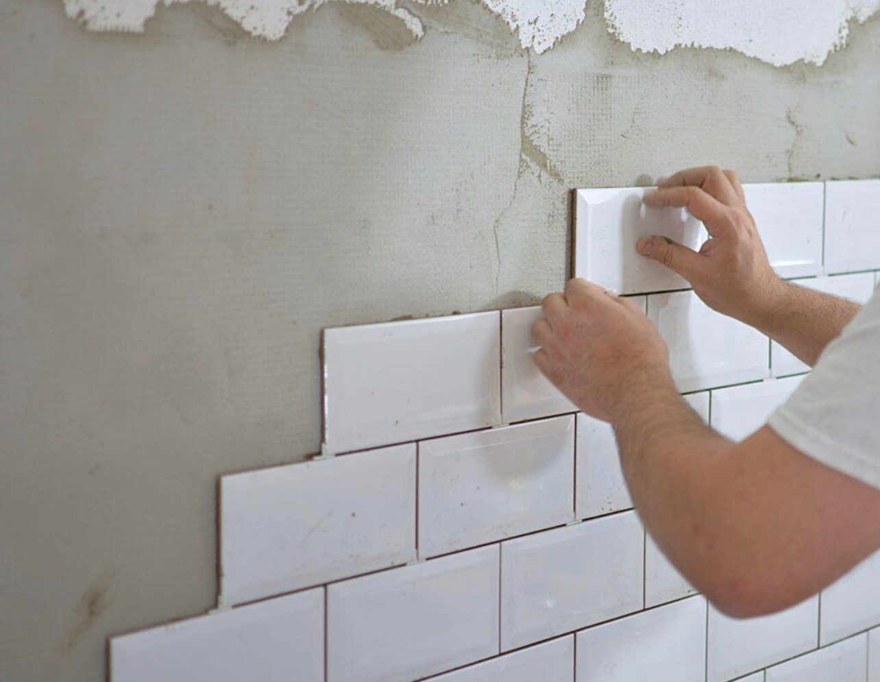 Układanie białych, prostokątnych płytek ściennych typu metro na szarej ścianie z widocznymi śladami zaprawy, etap prac wykończeniowych.