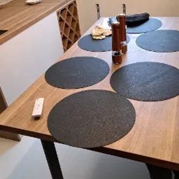 Drewniany blat stołu z czarnymi podkładkami, młynkami do pieprzu i soli oraz pilotem leżącym obok, w tle widoczny fragment blatu kuchennego z drewnianym wykończeniem i półkami na wino.