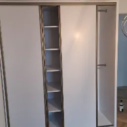 Biała szafa wnękowa z przesuwnymi drzwiami z widocznymi półkami i górnymi szafkami, na podłodze parkiet ułożony w jodełkę.