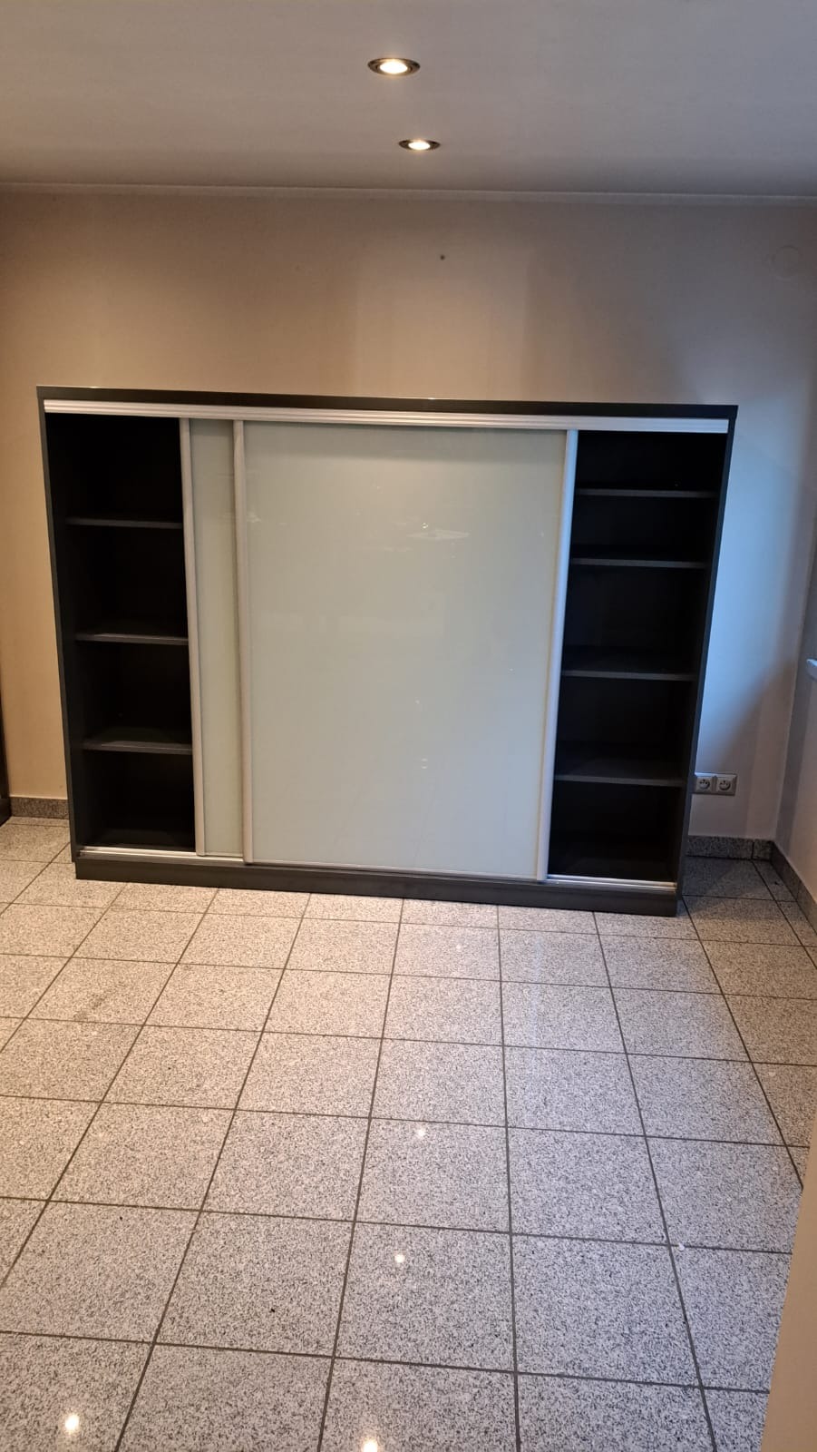 Duża szafa z przesuwnymi, mlecznymi drzwiami, z otwartymi półkami po bokach, na tle beżowej ściany i szarej podłogi z płytek.