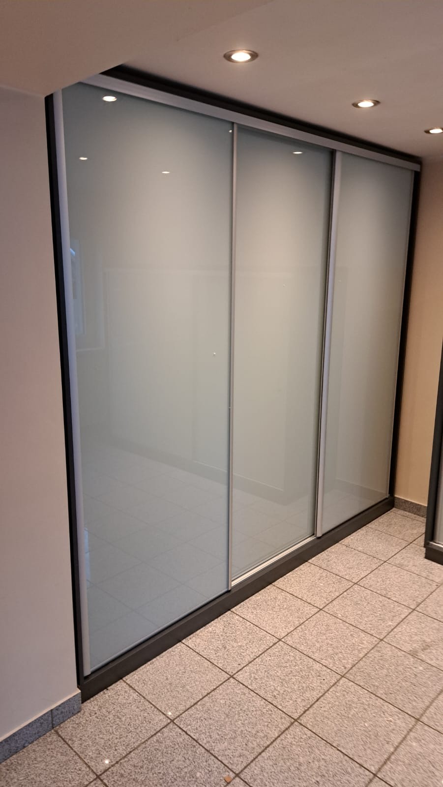 Trzyczęściowa szafa wnękowa z przesuwnymi, matowymi drzwiami w czarnej ramie, widoczna na tle jasnej podłogi z płytek.