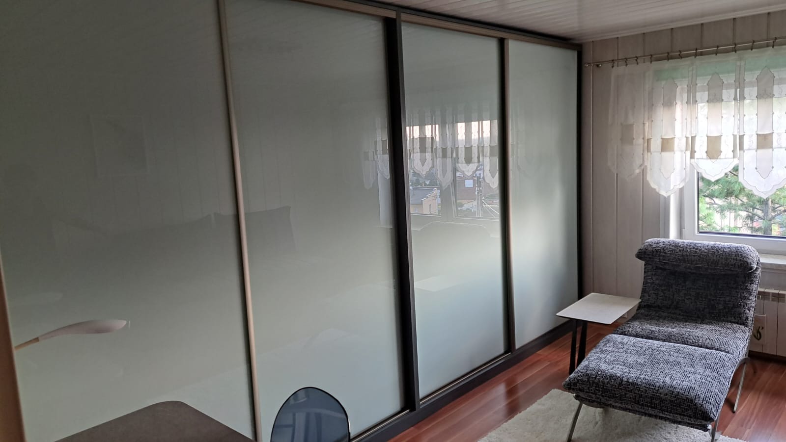 Duża szafa przesuwna z matowymi, jasnymi drzwiami w pokoju z drewnianą podłogą, widoczne odbicie wnętrza pokoju i okna z firanką.