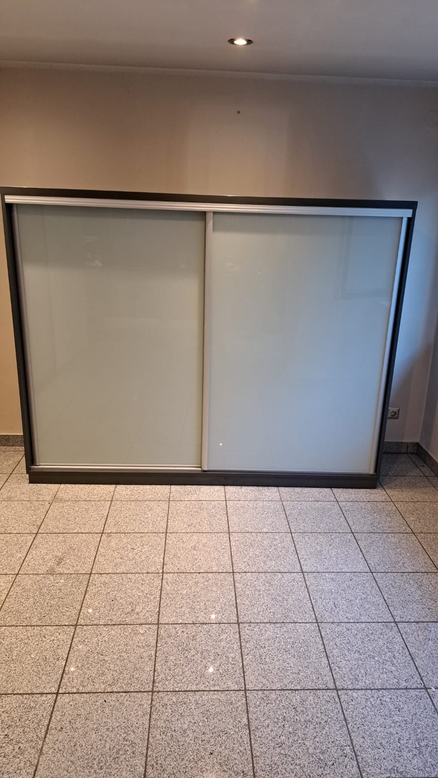 Szafa wnękowa z przesuwnymi, mlecznymi drzwiami w ciemnej ramie, widoczna na tle beżowej ściany i szarej podłogi z płytek.