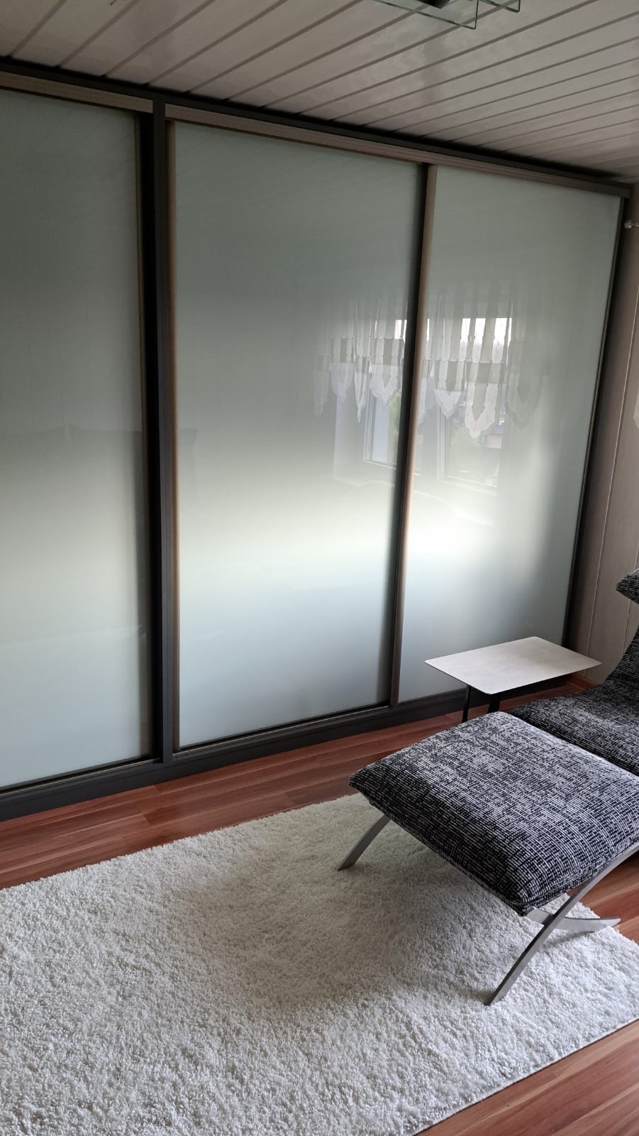 Trzydrzwiowa szafa przesuwna z matowego szkła w ciemnej ramie, uzupełniona pufą i stolikiem w jasnym wnętrzu z drewnianą podłogą i puszystym dywanem.