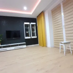 Nowoczesny salon z czarną ścianą akcentową, podświetlanym sufitem, białymi meblami RTV, wiszącymi witrynami i żółtymi zasłonami, uzupełniony o jasne rolety i mały biały stołek.