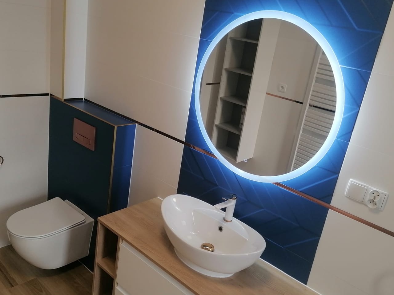 Nowoczesna łazienka z okrągłym lustrem LED, białą umywalką nablatową i toaletą podwieszaną, wykończona płytkami w odcieniach bieli i granatu z akcentami miedzianymi.