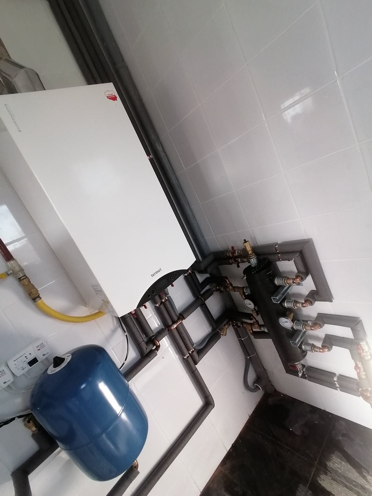 Biały piec gazowy Termet z niebieskim zbiornikiem wyrównawczym i rozbudowaną instalacją hydrauliczną z izolowanymi rurami, zamontowany na ścianie z białych płytek.
