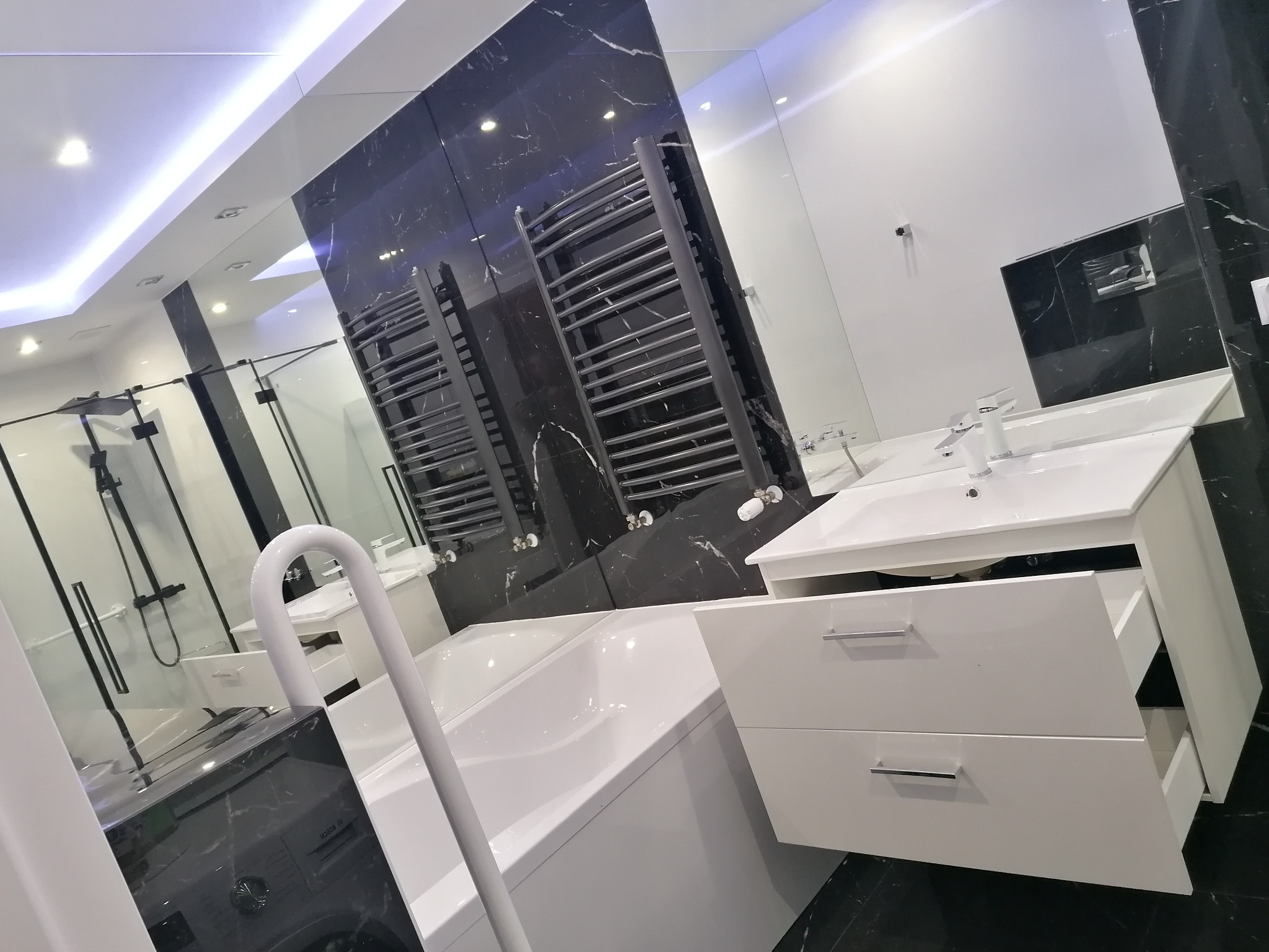 Nowoczesna łazienka z wanną, umywalką z szafką i grzejnikiem drabinkowym na tle czarnego marmuru.