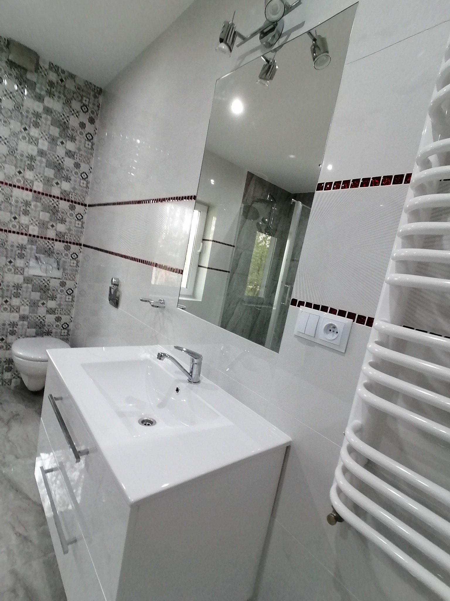 Nowoczesna łazienka z białą umywalką nablatową, grzejnikiem drabinkowym i lustrem z oświetleniem punktowym. Dekoracyjne płytki na ścianie i podłodze.