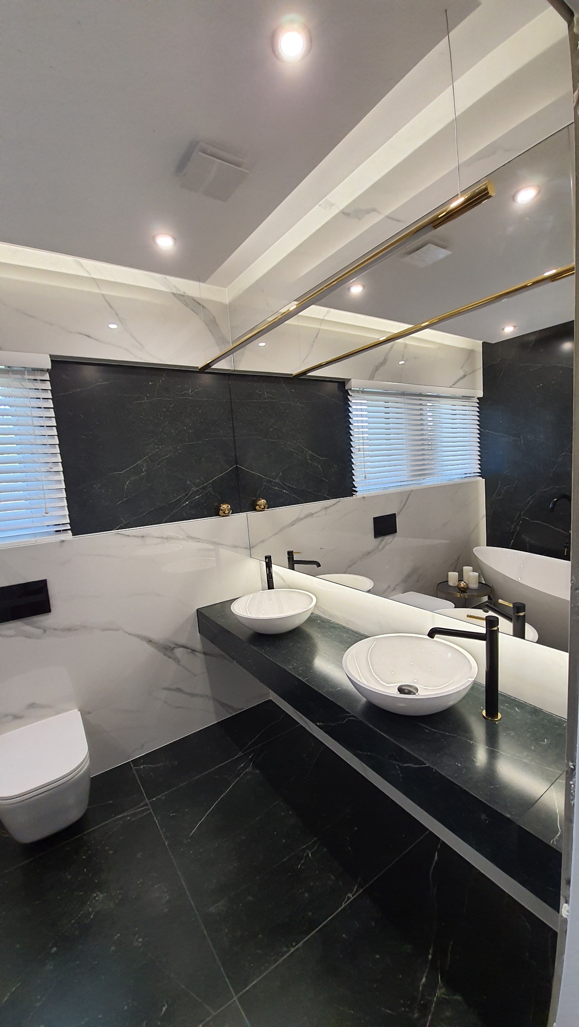 Nowoczesna łazienka z dwoma okrągłymi umywalkami nablatowymi, czarnymi bateriami, marmurowymi płytkami na ścianach i podłodze, oraz lustrem z złotymi akcentami, oświetlona punktowymi lampami...