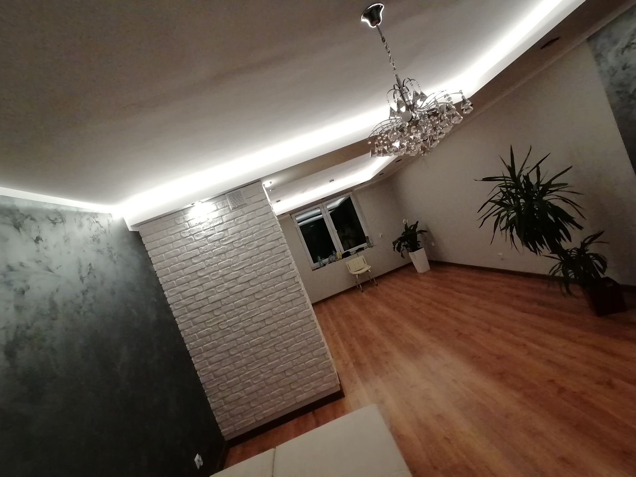 Nowoczesne wnętrze z drewnianą podłogą, ścianą z cegły pomalowanej na biało oraz dekoracyjną ścianą w odcieniach szarości, oświetlone listwą LED na suficie i kryształowym żyrandolem. Widoczne...