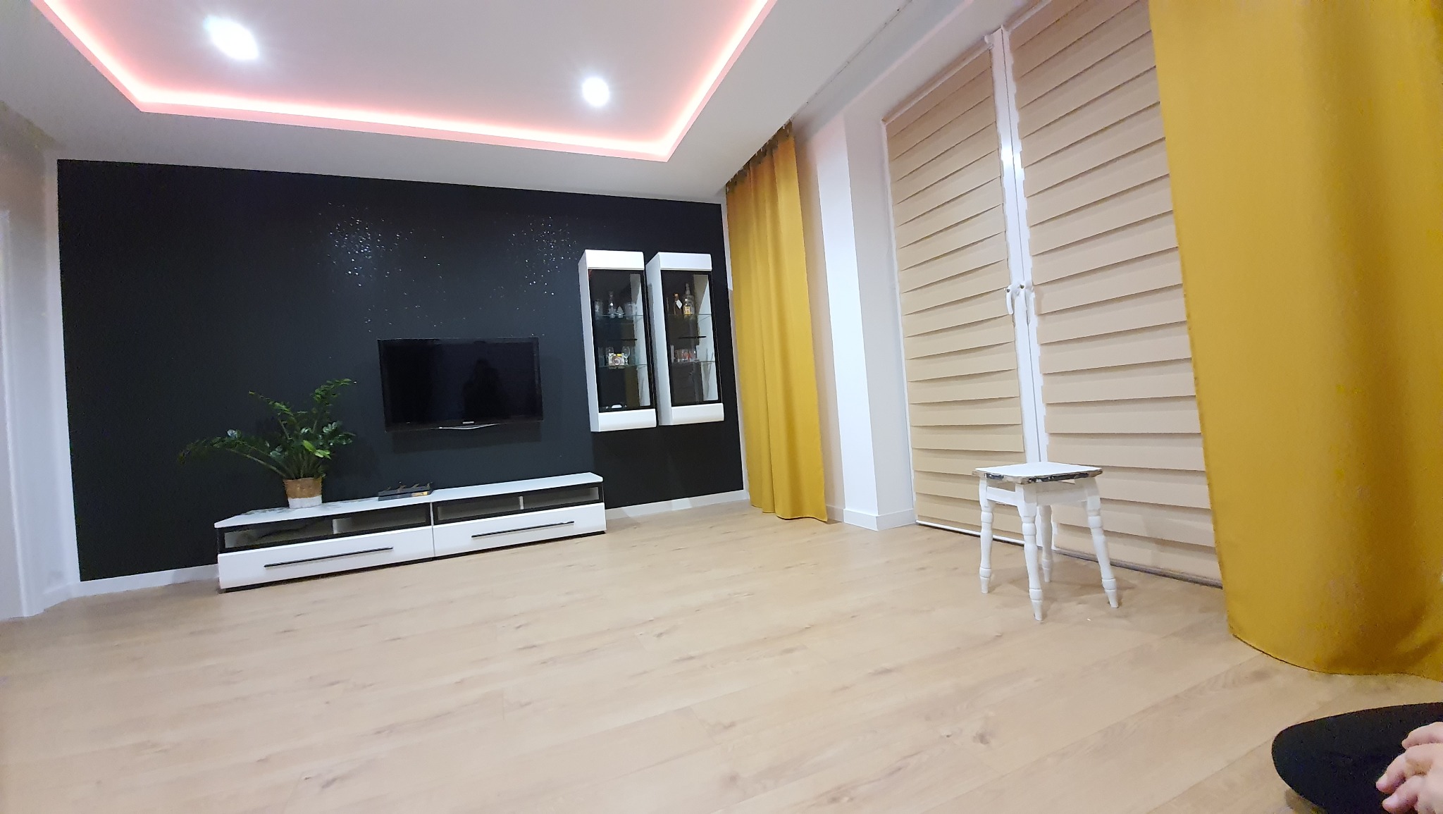 Nowoczesny salon z czarną ścianą akcentową, podświetlanym sufitem, białymi meblami RTV, wiszącymi witrynami i żółtymi zasłonami, uzupełniony o jasne rolety i mały biały stołek.