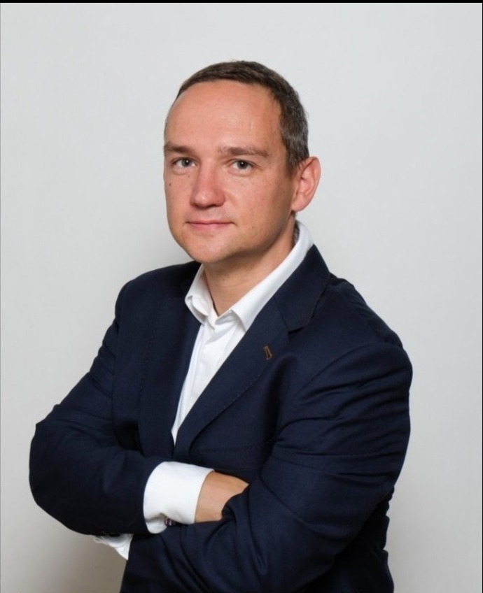 Portret mężczyzny w granatowym garniturze i białej koszuli, z założonymi rękami na tle jasnoszarego tła.