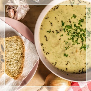 Kremowa zupa w różowym talerzu, posypana zieloną pietruszką, obok kromka chleba na serwetce, czosnek i cebula w tle.